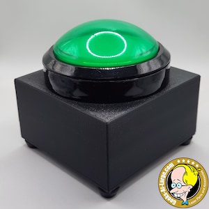 Op de afbeelding: Een grote, zwarte drukknop met een glanzende, groene koepel. De knop staat op een zwarte, vierkante basis met kleine voetjes. Het ontwerp is eenvoudig en modern, geschikt voor diverse toepassingen.