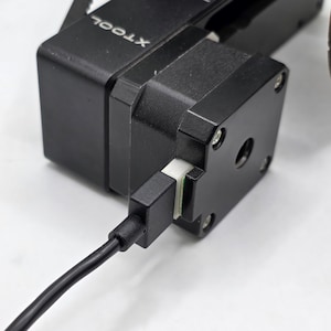 May include: A black XTOOL stepper motor component featuring a square housing with visible silver screws and a central circular opening. A black electrical cable is plugged into a white connector port on the side of the motor unit.