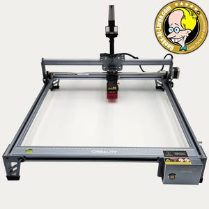 May include: A gray laser engraving machine with a red laser head and the word "CREALITY" printed on the frame. The machine has a rectangular frame and a control panel. The logo for "Geeks-R-Large.com" is in the upper right corner.