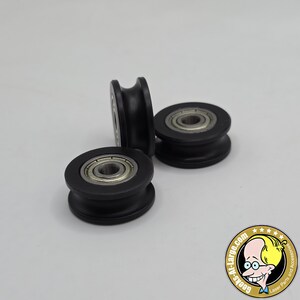 Puede incluir: Tres poleas negras con ranura en V con rodamientos de bolas plateados. Las ruedas están hechas de un material oscuro y mate y están diseñadas para sistemas mecánicos. La imagen está tomada sobre un fondo blanco.