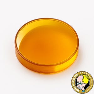 May include: A round, transparent orange lens. The lens has a smooth surface and a slightly raised edge. The image is on a white background. The lens is likely a component for laser parts and accessories.