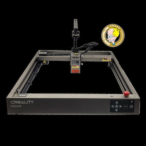 May include: A grey Creality Falcon2 laser engraver with a red emergency stop button. The machine has a rectangular frame and a central laser head. The control panel features directional buttons and a start/stop button. A sticker with a cartoon character is in the upper right corner.
