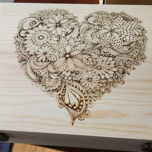 Könnte beinhalten: Holzkiste mit einem detaillierten herzförmigen Blumendesign. Das Design ist in das helle Holz eingebrannt und zeigt verschiedene Blumen- und Blättermuster. Die Kiste hat einen Metallverschluss und Scharniere.