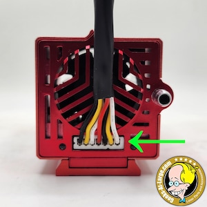 Può includere: Primo piano di un modulo laser rosso con un cavo nero e fili colorati. È visibile un connettore bianco, con una freccia verde che lo indica. Il modulo ha una ventola e un logo in basso a destra.