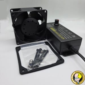 May include: Black computer fan with a power adapter and a bag of screws. The power adapter has a dial and a label that reads "ADAPTER POWER". A black square frame and screws are in a clear plastic bag.