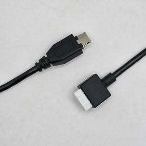 May include: A black electronic cable featuring a standard USB-type connector on one end and a white multi-pin rectangular connector housing on the other end. The cable is smooth, flexible, and black, connecting two distinct hardware interface components.