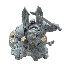 May include: A gray miniature figure of a warrior in ornate armor, wielding a sword and a weapon. The figure has large, feathered wings and a helmet. The miniature is on a textured base, likely for tabletop gaming.