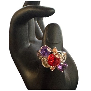 May include: A silver ring with a cluster of colorful gemstones. The ring features a large red stone surrounded by smaller purple and pink stones.
