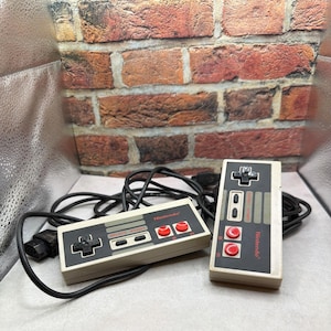May include: Two vintage Nintendo controllers with black directional pads, gray accents, and red buttons. The controllers are connected by black cords. The word "Nintendo" is printed on the controllers.