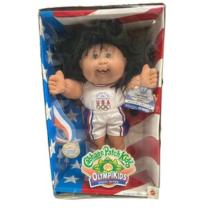May include: A Cabbage Patch Kids Olympikids doll with dark hair, wearing a white basketball jersey and shorts with USA and Olympic ring logos. The doll is inside a box decorated with a red, white, and blue American flag pattern and stars.