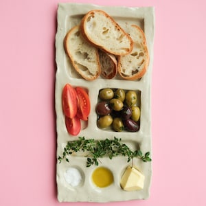 Puede incluir: Una bandeja de servicio rectangular de cerámica gris claro con múltiples compartimentos que contienen rebanadas de baguette, gajos de tomate rojo, aceitunas verdes y negras, hierbas frescas, sal, aceite de oliva y un cubo de mantequilla.