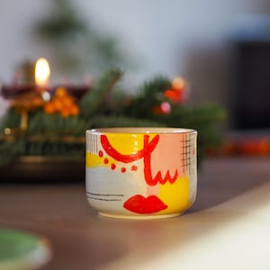 Pode incluir: Uma chávena de cerâmica com um design abstrato pintado à mão com formas vibrantes em vermelho, amarelo e rosa suave. O exterior inclui detalhes de linhas pretas, um padrão de grelha e um motivo de lábios vermelhos sobre um fundo cinzento.