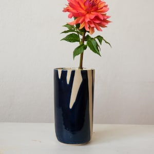 Può includere: Un vaso cilindrico in ceramica caratterizzato da una smaltatura blu navy scuro lucida con motivi a goccia verticali color crema. Un singolo fiore di dalia arancione e rosa vivace con foglie verdi è posizionato all'interno del vaso.