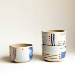 Puede incluir: Tres cuencos de cerámica con base color crema, decorados con líneas verticales y horizontales azules dibujadas a mano, parches rectangulares azul claro y pequeños puntos rojos. Un cuenco está solo, mientras que dos están apilados verticalmente.