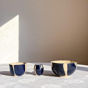 Könnte beinhalten: Drei Keramikgefäße mit einer glänzenden dunkelblauen Glasur und vertikalen cremefarbenen Streifen. Das Set besteht aus einer kleinen Schale, einem schmalen zylindrischen Becher und einer größeren Schale mit breitem Rand, alle handbemalt.