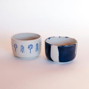 Pode incluir: Dois pequenos recipientes de cerâmica. O recipiente à esquerda é de cor branca suja com um padrão repetitivo de árvores desenhadas à mão em azul. O recipiente à direita apresenta um esmalte azul escuro brilhante com uma faixa vertical.