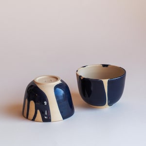Könnte beinhalten: Zwei Keramikschalen in Marineblau und Creme. Die Schalen haben eine abgerundete Form mit einem kontrastierenden Design. Eine Schale ist umgedreht und zeigt die Unterseite. Die andere Schale steht aufrecht.