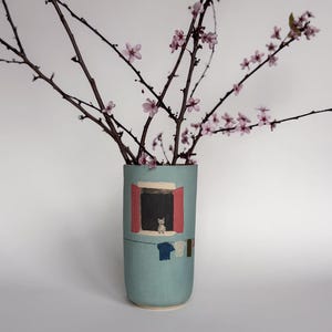 Könnte beinhalten: Hellblaue Keramikvase mit einer gemalten Szene eines Fensters mit einer Katze im Inneren und Wäsche, die draußen hängt. Die Vase enthält Zweige mit rosa Blüten. Die Vase ist etwa 20 cm hoch.