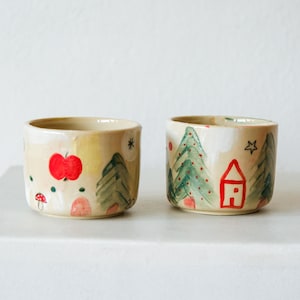 Puede incluir: Dos tazas de cerámica cilíndricas pequeñas con un esmalte de color crema. Las tazas presentan ilustraciones pintadas a mano que incluyen pinos verdes, una manzana roja brillante, una casa roja, una seta pequeña y varias estrellas y puntos.