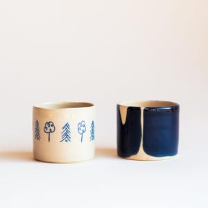 Könnte beinhalten: Zwei zylindrische Keramikbecher. Einer ist beige mit blauen Baum- und Blumenmustern. Der andere ist beige mit einer dunkelblauen Glasur auf der oberen Hälfte. Beide Becher haben ein schlichtes, modernes Design.
