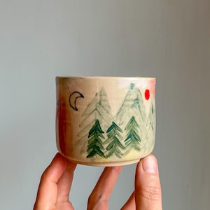 Puede incluir: Una pequeña taza de cerámica beige con una escena de bosque pintada a mano. El diseño incluye árboles verdes, montañas, un sol rojo y una luna creciente negra. La taza está sostenida en una mano.
