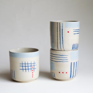 Op de afbeelding: Drie keramische bekers met een crèmekleurige basis, voorzien van handgeschilderde blauwe lijnpatronen, rasterontwerpen en lichtblauwe rechthoekige accenten. Elke beker is versierd met kleine, felrode stippen in verticale of horizontale lijnen.