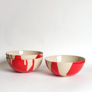 Puede incluir: Tres cuencos de cerámica con una base de color crema y rayas y goteos rojos irregulares. Dos cuencos están apilados a la izquierda, mientras que uno se encuentra a la derecha, todos con un acabado liso y brillante y bordes redondeados.