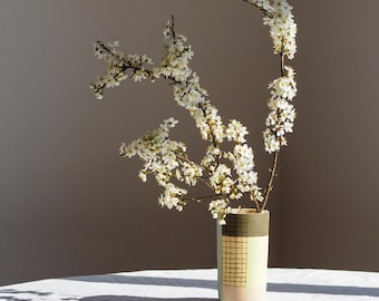 May include: A ceramic vase with a geometric design holds branches of white flowering blossoms. The vase has a light green, beige, and dark green color scheme. The vase is on a white tablecloth.