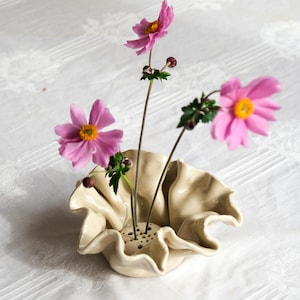Könnte beinhalten: Ein cremefarbener Blumenigel aus Keramik mit gewelltem, gerüschtem Rand und einem flachen Boden mit mehreren kleinen Löchern für Stiele. Drei rosa Anemonenblüten mit gelben Mitten und grünen Stielen stecken in den Löchern des Keramikobjekts.