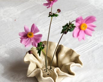May include: A cream-colored ceramic flower frog with a ruffled, wavy edge and a flat base featuring multiple small holes for stems. Three pink anemone flowers with yellow centers and green stems are inserted into the holes of the ceramic piece.