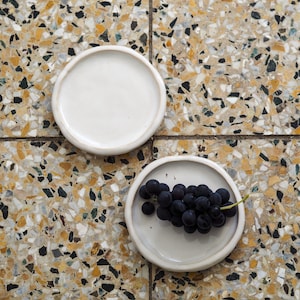 Könnte beinhalten: Zwei kleine, runde, weiße Keramikplatten auf einer gefleckten Fliesenoberfläche. Eine Platte ist leer, die andere enthält eine Traube dunkler, violetter Trauben. Die Teller sind schlicht und modern gestaltet.