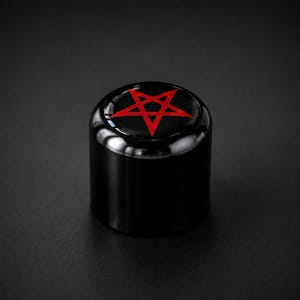 Könnte beinhalten: Ein schwarzes zylindrisches Objekt mit glänzender Oberfläche. Auf der Oberseite befindet sich ein rotes Pentagramm-Design. Das Objekt ist vor einem dunklen Hintergrund platziert, der seine Form und die kontrastierenden Farben des Designs hervorhebt.