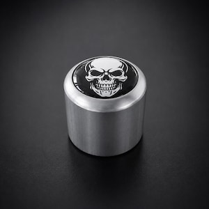 Könnte beinhalten: Zylindrisches Objekt aus gebürstetem Metall mit einem schwarz-weißen Totenkopf-Design auf der Oberseite. Der Totenkopf hat detaillierte Augenhöhlen, eine Nasenhöhle und ein vollständiges Gebiss. Es ist wahrscheinlich ein dekoratives Element oder ein Knopf.