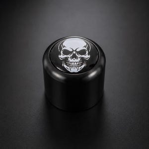 Pode incluir: Uma rolha de garrafa cilíndrica preta com um desenho de caveira branca na parte superior. A caveira tem órbitas oculares detalhadas, uma cavidade nasal e uma dentição completa. A rolha é feita de um material brilhante e reflexivo.