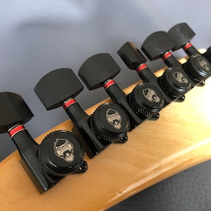 Puede incluir: Primer plano de seis clavijas de afinación de guitarra negras con detalles rojos. Cada clavija tiene una perilla negra con un logotipo de cabeza de gorila plateado.