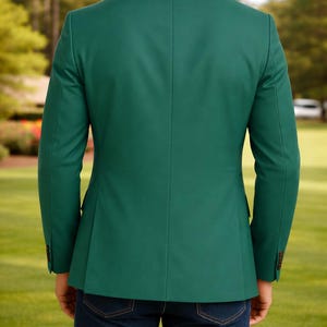 May include: A solid emerald green blazer with a two-button closure, worn with dark blue denim jeans. The blazer has a tailored fit and long sleeves. The background shows a green lawn and trees.