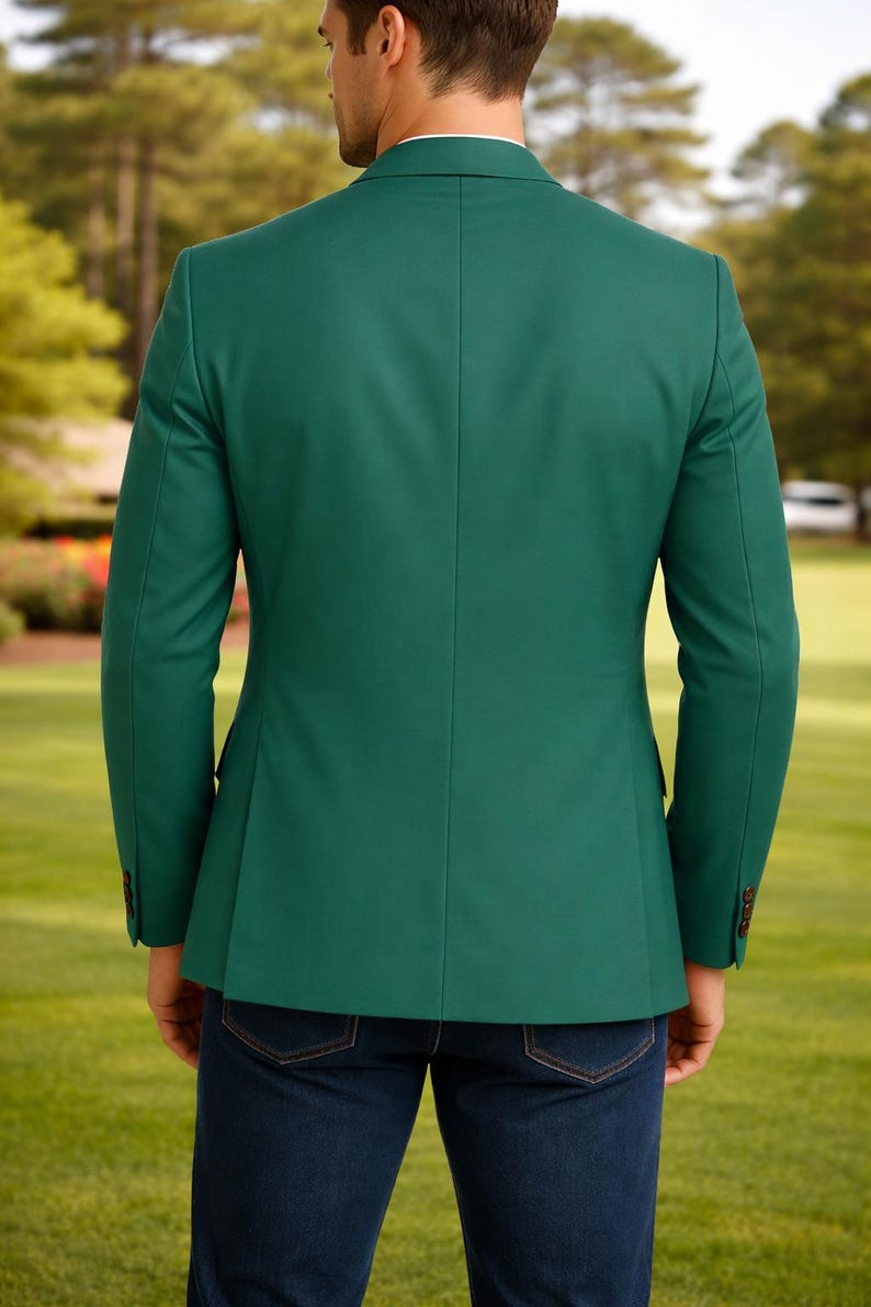 May include: A solid emerald green blazer with a two-button closure, worn with dark blue denim jeans. The blazer has a tailored fit and long sleeves. The background shows a green lawn and trees.