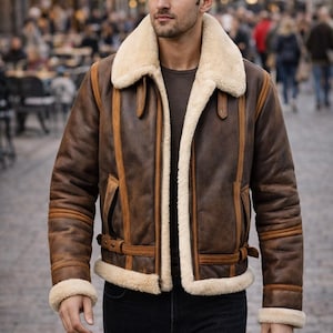 Könnte beinhalten: Braune Lederjacke mit cremefarbenem Shearling-Kragen und -Futter. Die Jacke hat einen Reißverschluss, Seitentaschen und einen Schnallenriemen. Die Ärmel haben Shearling-Manschetten. Die Person trägt eine schwarze Jeans.