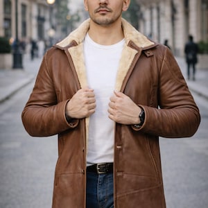 Könnte beinhalten: Ein brauner Ledermantel mit Schafsfellkragen und -futter. Der Mantel ist zugeknöpft und wird über einem weißen T-Shirt und einer blauen Jeans getragen. Ein schwarzer Gürtel mit silberner Schnalle ist sichtbar.