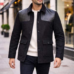May include: A black jacket with leather shoulder patches and a collar, worn over a white sweater. The jacket has two front pockets and button closures. The person is wearing dark blue jeans.
