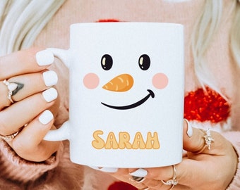May include: A white ceramic mug with a snowman face and the name "SARAH" printed in orange. The mug is being held by a person wearing a pink sweater with a Christmas pattern.