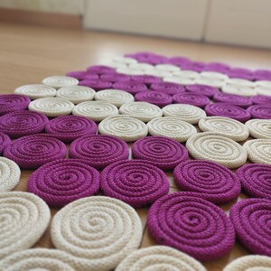 May include: A close-up of a woven rug made of thick rope. The rug is made up of a series of circles in alternating colours of cream and purple.