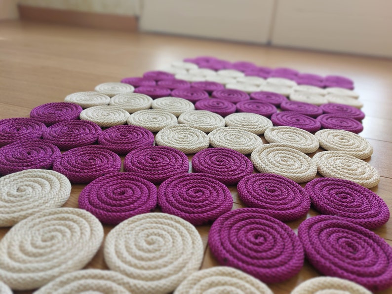 May include: A close-up of a woven rug made of thick rope. The rug is made up of a series of circles in alternating colours of cream and purple.