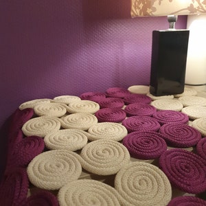 May include: A close-up of a beige and burgundy circular rug made of woven rope. The rug is made up of many circles, each with a different size and pattern. The rug is on a table with a lamp in the background.