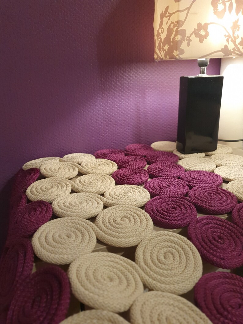 May include: A close-up of a beige and burgundy circular rug made of woven rope. The rug is made up of many circles, each with a different size and pattern. The rug is on a table with a lamp in the background.