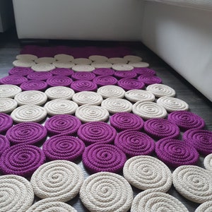May include: A handmade rug featuring a circular pattern in shades of purple and beige. The rug is crafted from braided rope and has a contemporary, minimalist design.