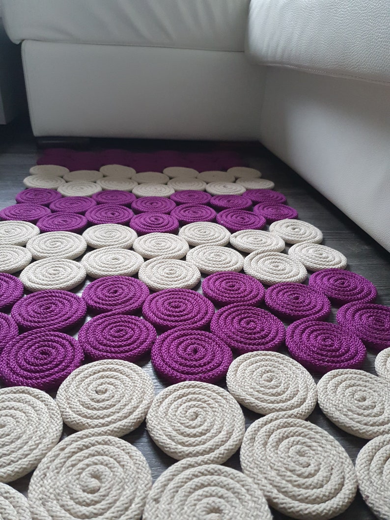 May include: A handmade rug featuring a circular pattern in shades of purple and beige. The rug is crafted from braided rope and has a contemporary, minimalist design.