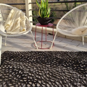 Peut inclure: Un tapis d'extérieur noir à motifs avec un motif de boucles répétées. Deux chaises blanches tissées avec des couvertures crème sont de chaque côté d'une petite table d'appoint rouge. Une plante en pot est sur la table, sur un balcon.
