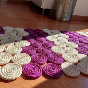 May include: A close-up of a circular rug made of woven rope. The rug is made up of alternating circles of cream and purple.