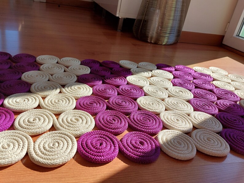 May include: A close-up of a circular rug made of woven rope. The rug is made up of alternating circles of cream and purple.